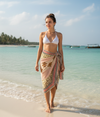 Woman in a white bikini top and cotton beach wrap standing on a beach with clear blue water and sky.