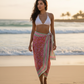 Woman in a white bikini top and pink sarong standing on a beach at sunset.