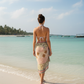 Woman in a floral dress standing on a beach with clear blue water and sky.