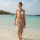 Woman in a white bikini top and cotton beach wrap standing on a beach with clear blue water and sky.